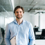 Homme souriant en chemise bleue dans un bureau moderne, tenant un classeur.