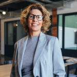 Femme blonde avec lunettes et blazer bleu clair dans un bureau moderne.
